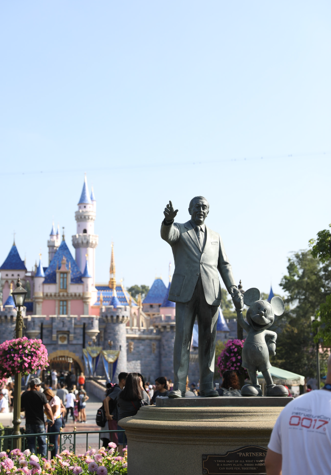 An image of the castle at Disneyland and the statue of Walt holding Mickys hand. In the foreground is a man with an instructure con 2017 shirt on